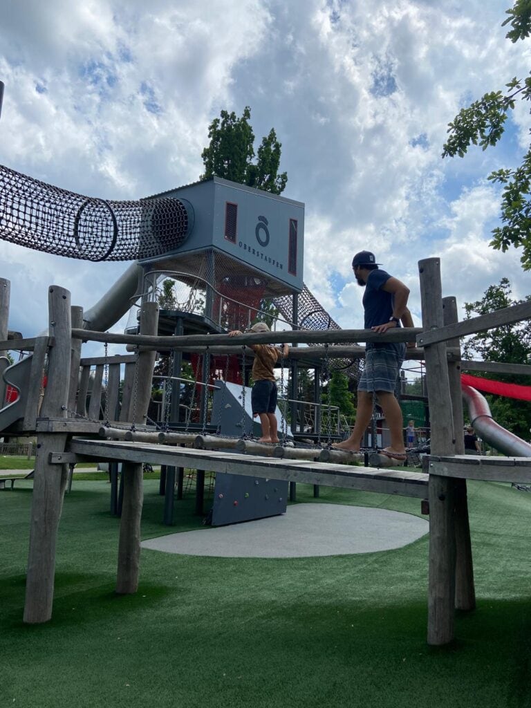 Ein hoher Spielturm auf einem gepflegten Rasen und davor eine Hängebrücke, auf der Vater und Sohn gehen.