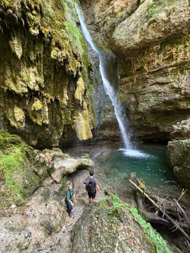 Eine Familie wandert am Hinanger Wasserfall und das Wasser stürzt von hoch oben in ein Felsenbecken